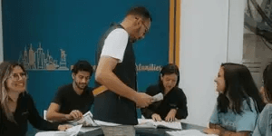 cinco pessoas em uma sala, quadro delas sentadas sobre uma mesa com uma pessoa em pé dando aula